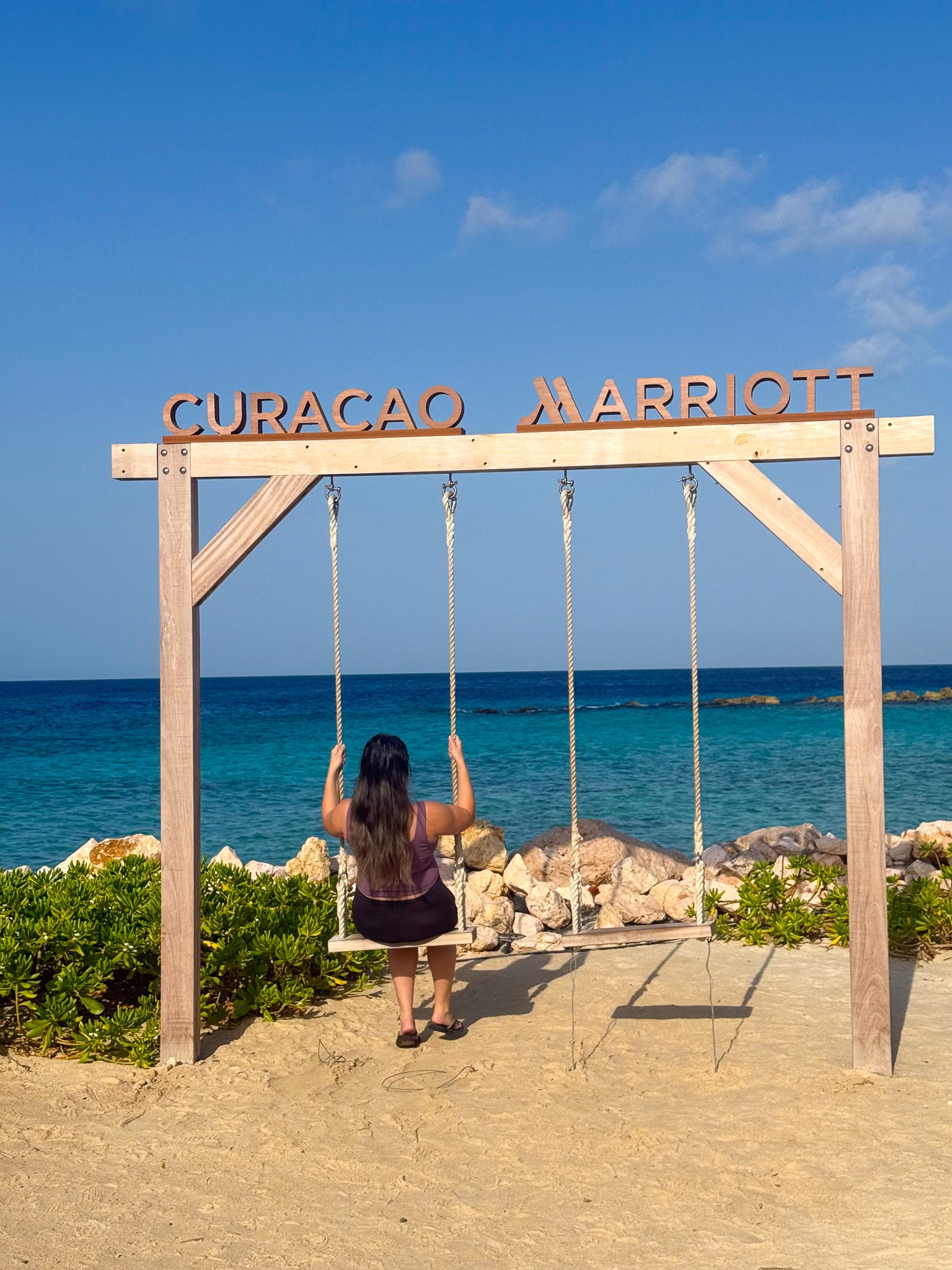 Back view of a woman seated on a wooden swing facing the turquoise sea with a Curacao Marriott sign overhead.