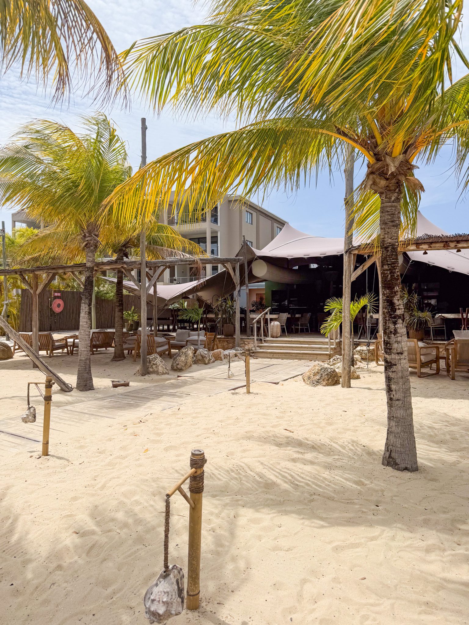 Beachside cafe with palm trees, white canopies, and wooden seating on sand.